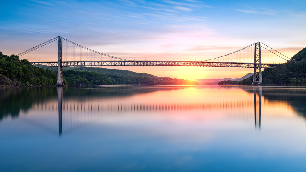 A bridge spanning a wide, calm river, with the sun rising behind it