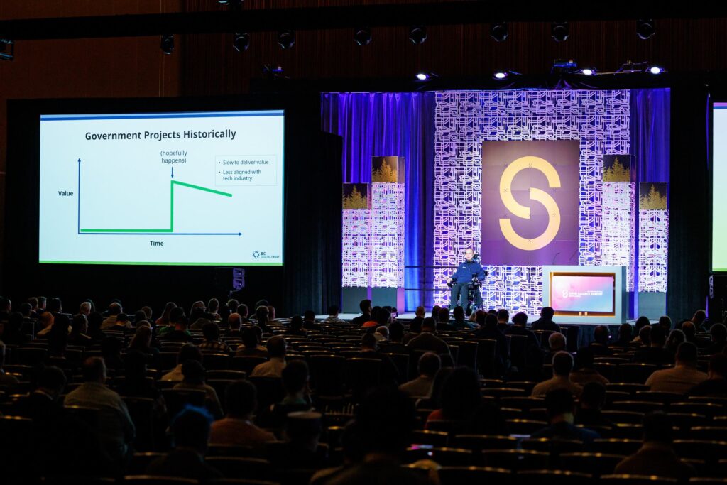 A picture of John Jordan on stage presenting his keynote as Open Source Summit North America, with a crowd in darkness in the foreground